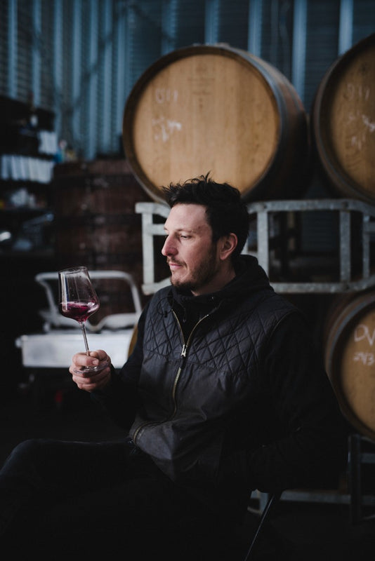 Man seated in front of wine barrels swirls wine in a glass